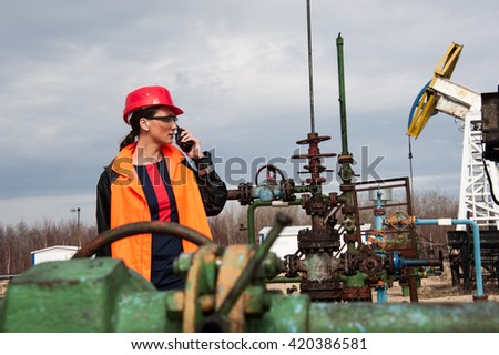 Woman engineer in the oil field talking on the radio wearing red helmet and orange work clothes. Industrial site background.