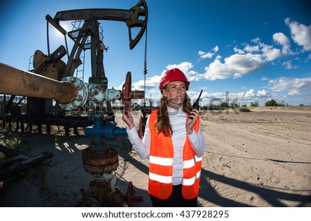Woman engineer in the oil field talking on the radio wearing red helmet and orange work clothes. Industrial site background.