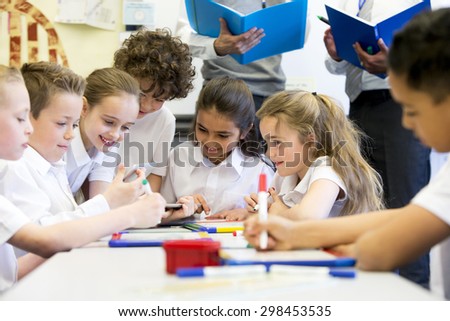 A group of school children can be seen working on digital tablets and whiteboards, they are all working happily. Two unrecognizable teachers can be seen in the background.