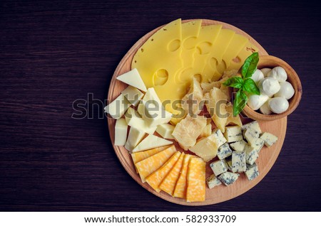 Cheese plate: Parmesan, cheddar, gouda, mozzarella and other with basil on wooden board on dark background with place for text. Tasty appetizers. Top view. Copy space.