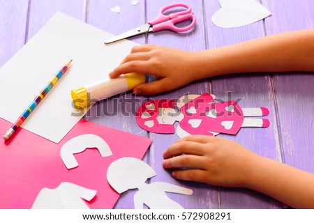 Small child made an angel doll of cardboard. Childrens hands on a wooden table. Craft supplies for a fun kids activity. Valentine's day crafts idea for kids in home or kindergarten