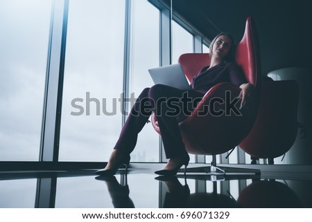 Tired and relaxed boss woman with laptop is sprawling in bent red armchair in office chill out area and dreamy looking up, with copy space place for logo, your text or other advertising