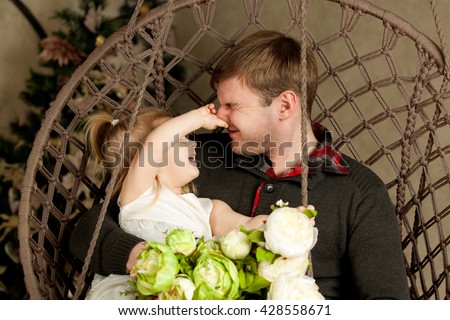 Daughter plays with dad. Babe caught father's nose and man's eyes were closed. Funny family photo. Blonde girl in a white dress sitting on dad's lap