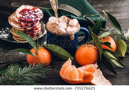 A mug with cocoa and melted marshmallows. Hot chocolate. Mandarins and pancakes with jam on a wooden table.