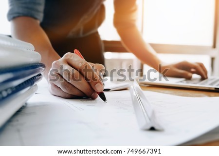 Young engineers holding a red pen pointing to a building on a blueprint and using a computer laptop to write a presentation for the meeting in working site.