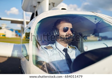Portrait of cool young adult pilot sitting in private air plane ready to take off. Bright summer day, handsome male model