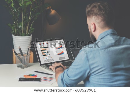 Back view. Young bearded businessman in denim shirt is sitting in office at table and is using laptop with charts, graphs and diagrams on screen. On table is smartphone and stationery. Man working.