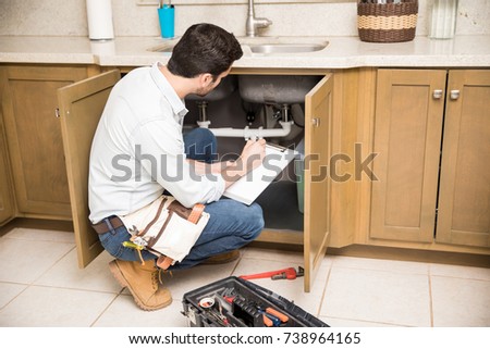 Rear view of a male plumber writing a repair order while crouching in front of a kitchen sink