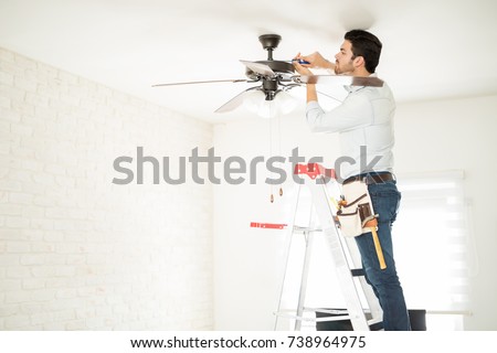 Profile view of a male electrician stepping on a ladder and installing a ceiling fan in a house