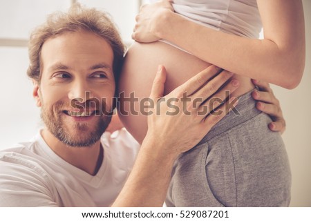 Handsome man is listening to his beautiful pregnant wife's tummy and smiling