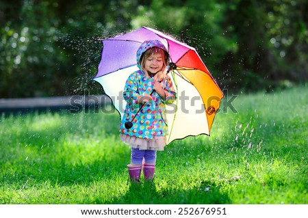 Funny cute toddler girl wearing waterproof coat with colorful umbrella playing in the garden by rainy and sunny day