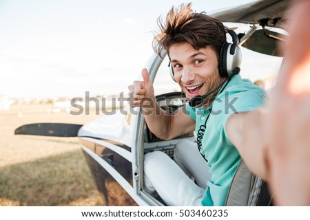 Cheerful excited young pilot sitting in cabin of small airplane and showing thumbs up