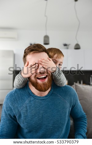 Photo of playful bearded father dressed in blue shirt playing with his little cute son who covering father's eyes indoors