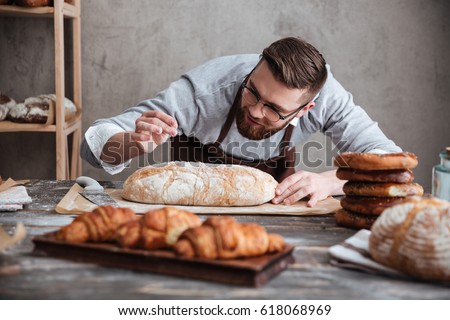 Picture of young concentrated man baker standing at bakery near bread. Looking aside.