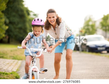 A laughing, smiling mother pushes her daughter forward on a warm summer's day as she teaches her how to ride her bicycle on a city sidewalk near a green park.