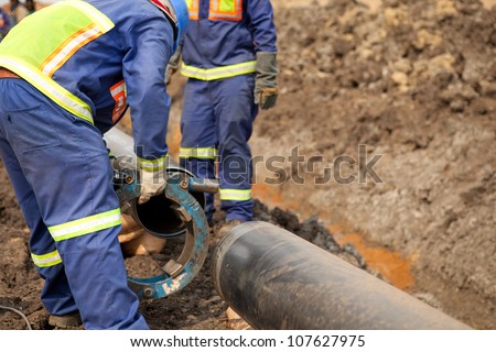 Male welder workers wearing protective high visibility clothing fixing and joining industrial construction oil and gas or water plumbing pipeline using an external pipe clamp outside on site