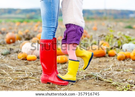 Legs of young woman and her little kid girl daugher in rainboots. Woman in red gum boots, child in yellow shoes. On pumpkin field, outdoors.