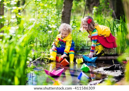 Children play with colorful paper boats in a small river on a sunny spring day. Kids playing exploring the nature. Brother and sister having fun at a forest stream. Boy and girl with toy boat and ship
