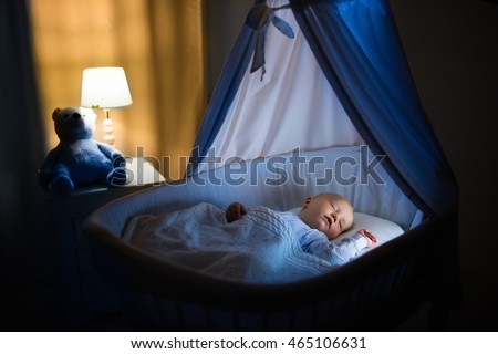 Adorable baby sleeping in blue bassinet with canopy at night. Little boy in pajamas taking a nap in dark room with crib, lamp and toy bear. Bed time for kids. Bedroom and nursery interior.