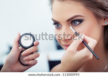 Make-up artist applying white eyeshadow in the corner of model's eye and holding a shell with eyeshadow on background, close up