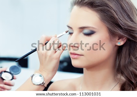 Make-up artist applying bright base color eyeshadow on model's eye and holding a shell with eyeshadow on background, close up