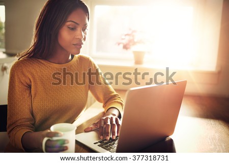 Serious Black adult single female sitting at table holding coffee cup and typing on laptop with light flare coming through window