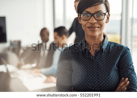 Beautiful young grinning professional Black woman in office with eyeglasses, folded arms and confident expression as other workers hold a meeting in background