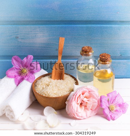 Spa setting. Sea salt in bowl and towels, aroma oil and flowers on white wooden background against blue wall. Selective focus. Place for text. Square image.