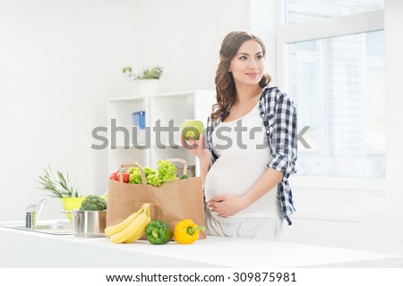 Beautiful pregnant woman in the kitchen with shopping bag and apple.