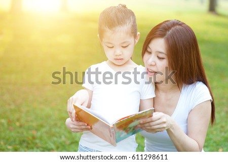 Asian mother and daughter are reading book and having fun in nature. Family outdoor fun, morning with sun flare.