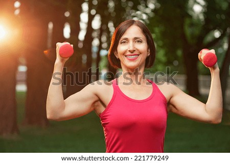 Portrait of cheerful aged woman in fitness wear exercising with red dumbbells in park.