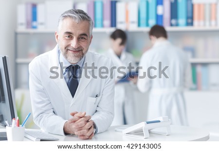 Smiling confident doctor at the reception desk, medical staff working on the background