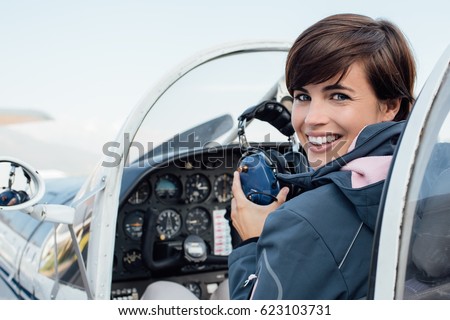Smiling female pilot in the light aircraft cockpit, she is holding aviator headset and looking at camera