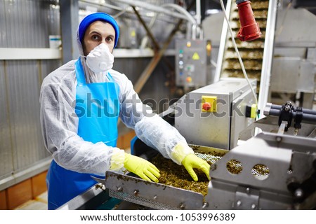 Young man in mask, gloves and coveralls looking at camera while working iwth processed seaweed in factory