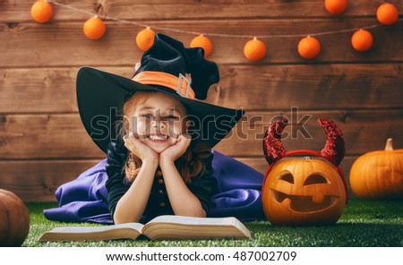 Happy Halloween! Cute cheerful little witch with a book of spells. Beautiful child girl in witch costume sitting on grass, reading and laughing.