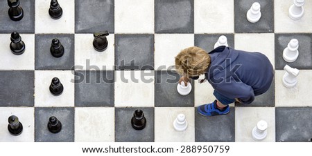 Young boy moving a white pawn during a chess game on an outdoor chess board, seen from above