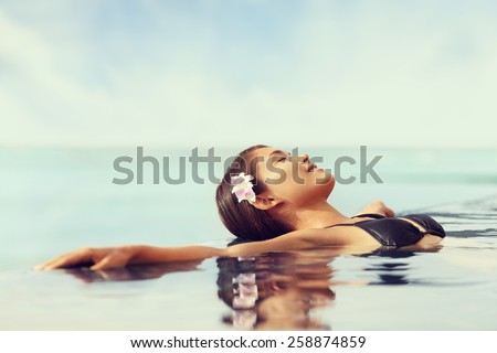 Luxury resort woman relaxing in infinity swim pool. Asian young adult lying down in swimming pool of beach resort for summer holidays or travel vacations.
