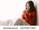 Small photo of Portrait of beautiful Asian woman relaxing with cup of hot cocoa or chocolate with marshmallow while sitting by the window. Happy girl enjoying warm beverage in winter day. Lazy day off concept.