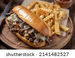 Small photo of Philly cheesesteak sandwich made with steak, cheese and onions on a hoagie roll with french fries on a wooden board