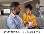 Small photo of Indian father and smiling son putting coin into piggy bank. Smiling boy sitting on father lap saving money in piggybank. Middle eastern dad teaching son to save money while putting coin in piggy bank.