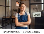 Small photo of Portrait of happy waitress standing at restaurant entrance. Portrait of young business woman attend new customers in her coffee shop. Smiling small business owner showing open sign in her shop.