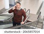Small photo of Smiling mature man standing in escalator while talking over smartphone. Successful business man in airport standing on escalator while doing a conversation on the phone. Mid adult man going to work.