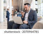 Small photo of Serious businessman sitting on couch in airport lounge using laptop and sending voice message with smartphone. Mature business man working on computer while using voice command recorder on phone.