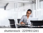 Mature businesswoman taking notes in notebook while using laptop at office. Mid adult black woman entrepreneur writing details on paper while working on laptop. Smiling african american business woman