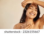 Happy young woman with bare shoulder applying serum on face with closed eyes. Beautiful hispanic young woman moisturizes her skin with serum isolated against beige background with copy space.