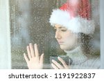 Sad and lonely woman looking through a window touching glass, with the hand in christmas time in a rainy day