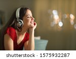 Happy woman wearing wireless headphones listening to music in the night sitting on a couch in the living room at home