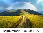 Amazing scene in summer mountains. Lush green grassy meadows in fantastic evening sunlight. Rural road and beautyful rainbow in dramatic sky. Landscape photography