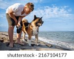 Small photo of Cheerful kind teenage guy with blond hair and comfortable leash in his hands plays and walks with his big fluffy multi-colored dog of Akina Inu breed, on sandy wild sea beach along Black Sea