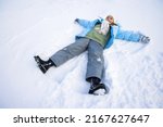 Above view of cute little girl making snow angel in fresh deep snow. Female child wearing ski clothes and skarf lying, waving arms and legs, making angel on snow in park. Concept of winter activities.